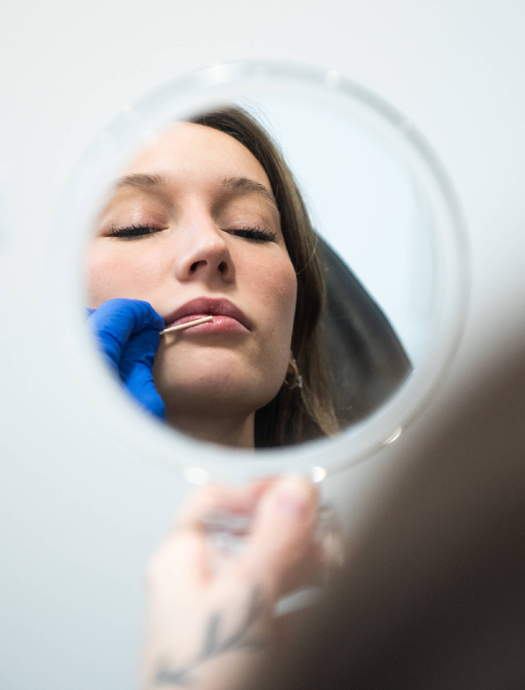 Patiente examinant ses lèvres dans un miroir après une injection d’acide hyaluronique dans un centre médico-esthétique. Traitement esthétique réalisé par un professionnel avec des gants bleus pour repulper et améliorer le volume naturel des lèvres.